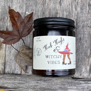 A dark candle jar sits on a vintage wood surface.  The label reads Thick Thighs & Witchy Vibes.  The graphic is anime inspired witch legs with black and orange striped stockings, topped with red witch hat.