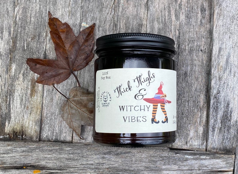 A dark candle jar sits on a vintage wood surface.  The label reads Thick Thighs & Witchy Vibes.  The graphic is anime inspired witch legs with black and orange striped stockings, topped with red witch hat.