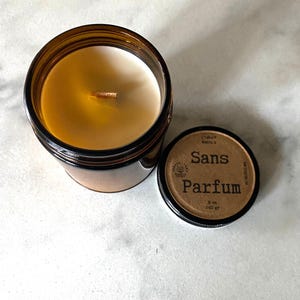 May include: A brown glass jar candle with a white wax interior and a wooden wick. The lid is off, revealing the candle's contents. The lid has the words "Sans Parfum" printed on it, along with other text. The candle is 8 oz / 240 gr.