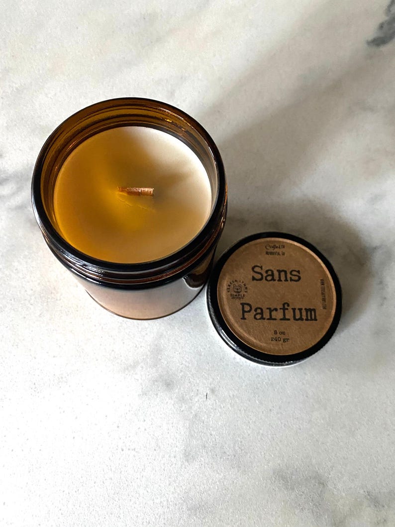 May include: A brown glass jar candle with a white wax interior and a wooden wick. The lid is off, revealing the candle's contents. The lid has the words "Sans Parfum" printed on it, along with other text. The candle is 8 oz / 240 gr.