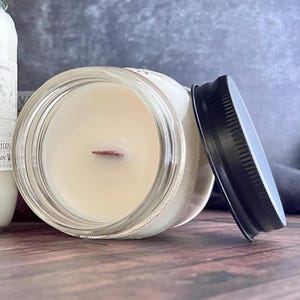 May include: A clear glass jar candle with a black lid. The candle has a light beige wax and a wooden wick. The lid is off to the side. The candle is on a wooden surface.