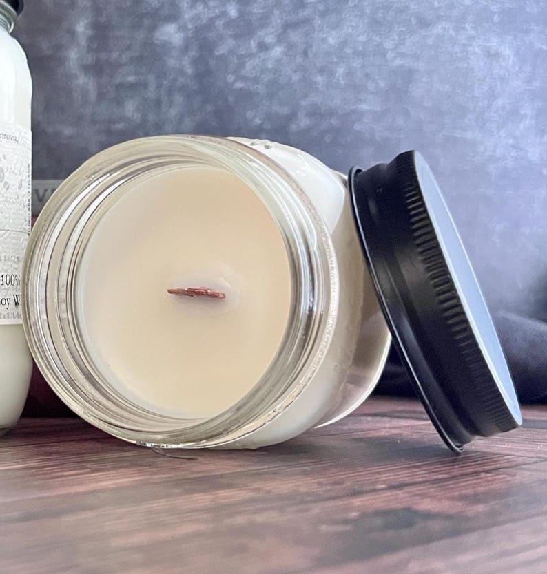 May include: A clear glass jar candle with a black lid. The candle has a light beige wax and a wooden wick. The lid is off to the side. The candle is on a wooden surface.