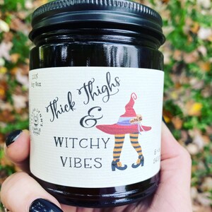 A hand with long black finger nails holds a dark candle jar.  The label reads Thick Thighs & Witchy Vibes.  The graphic is anime inspired witch legs with black and orange striped stockings, topped with red witch hat.  The background is a fall scene.