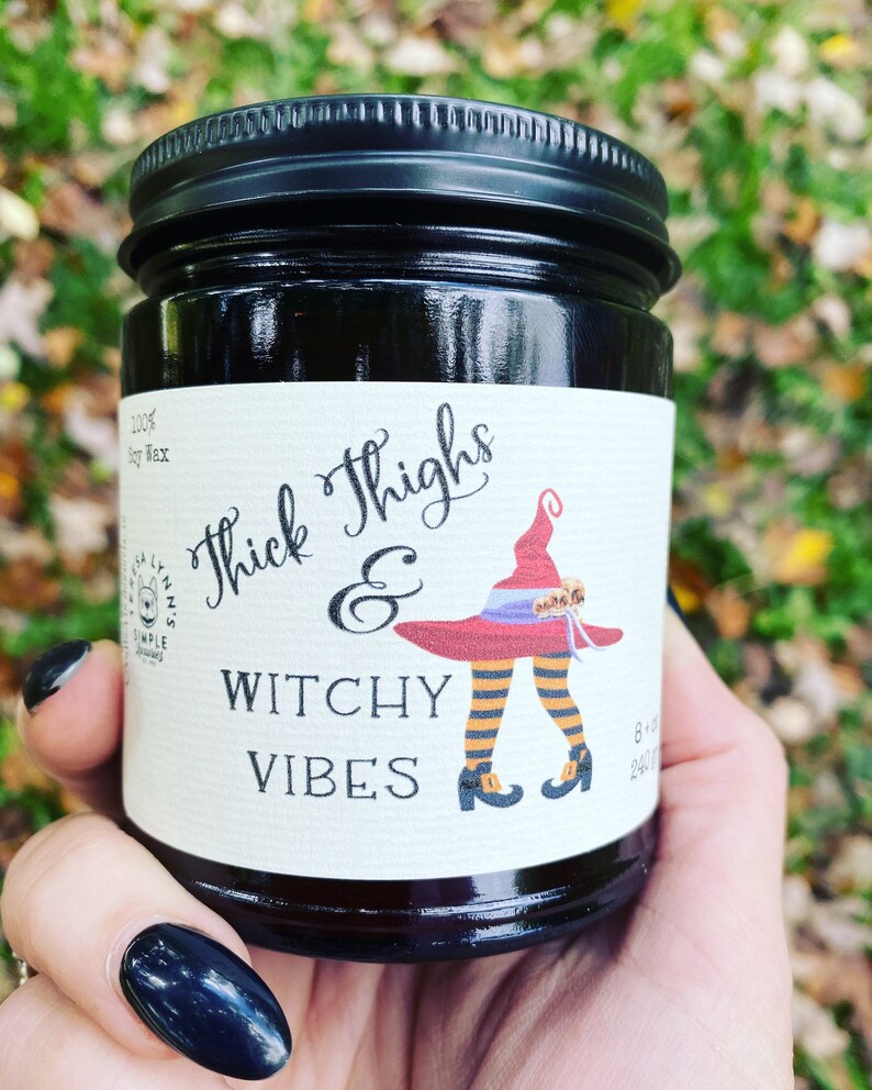A hand with long black finger nails holds a dark candle jar.  The label reads Thick Thighs & Witchy Vibes.  The graphic is anime inspired witch legs with black and orange striped stockings, topped with red witch hat.  The background is a fall scene.