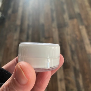 Op de afbeelding: Een kleine, cilindervormige witte plastic container met een doorzichtige basis en een plat wit deksel. De container is compact en heeft een glad, ondoorzichtig wit lichaam, ontworpen voor het bewaren van kleine hoeveelheden inhoud.
