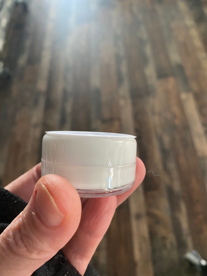 Op de afbeelding: Een kleine, cilindervormige witte plastic container met een doorzichtige basis en een plat wit deksel. De container is compact en heeft een glad, ondoorzichtig wit lichaam, ontworpen voor het bewaren van kleine hoeveelheden inhoud.