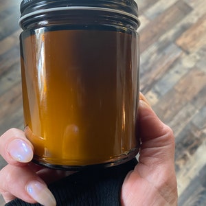 May include: A clear glass jar with a black lid. The jar is filled with a golden-brown substance. The jar is held by a person wearing a black long-sleeve shirt.