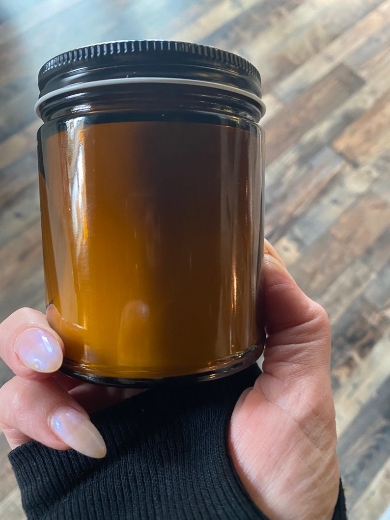 May include: A clear glass jar with a black lid. The jar is filled with a golden-brown substance. The jar is held by a person wearing a black long-sleeve shirt.