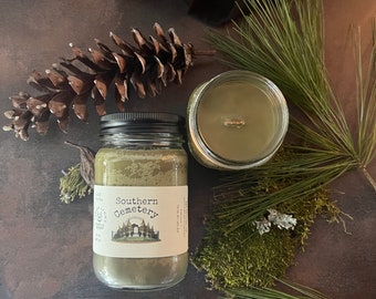 Pode incluir: Um frasco de vela de vidro verde com uma tampa preta e um rótulo branco que diz "Southern Cemetery". A vela está rodeada de agulhas de pinheiro e uma pinha.
