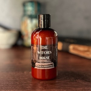 An Amber colored bottle of body wash labeled Witches House, crafted in Monrovia,In sits in from of a blurred background that includes a grimoire and mortal and pestle.