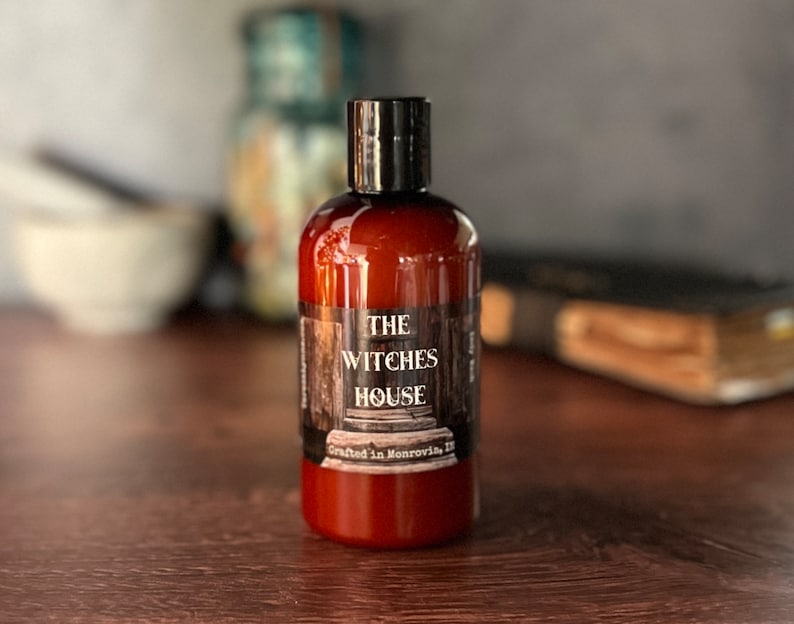 An Amber colored bottle of body wash labeled Witches House, crafted in Monrovia,In sits in from of a blurred background that includes a grimoire and mortal and pestle.