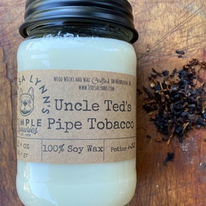 May include: A glass jar candle with a black lid. The label is brown with white text that reads "Uncle Ted's Pipe Tobacco" and "100% Soy Wax". There is a small illustration of a dog on the label.