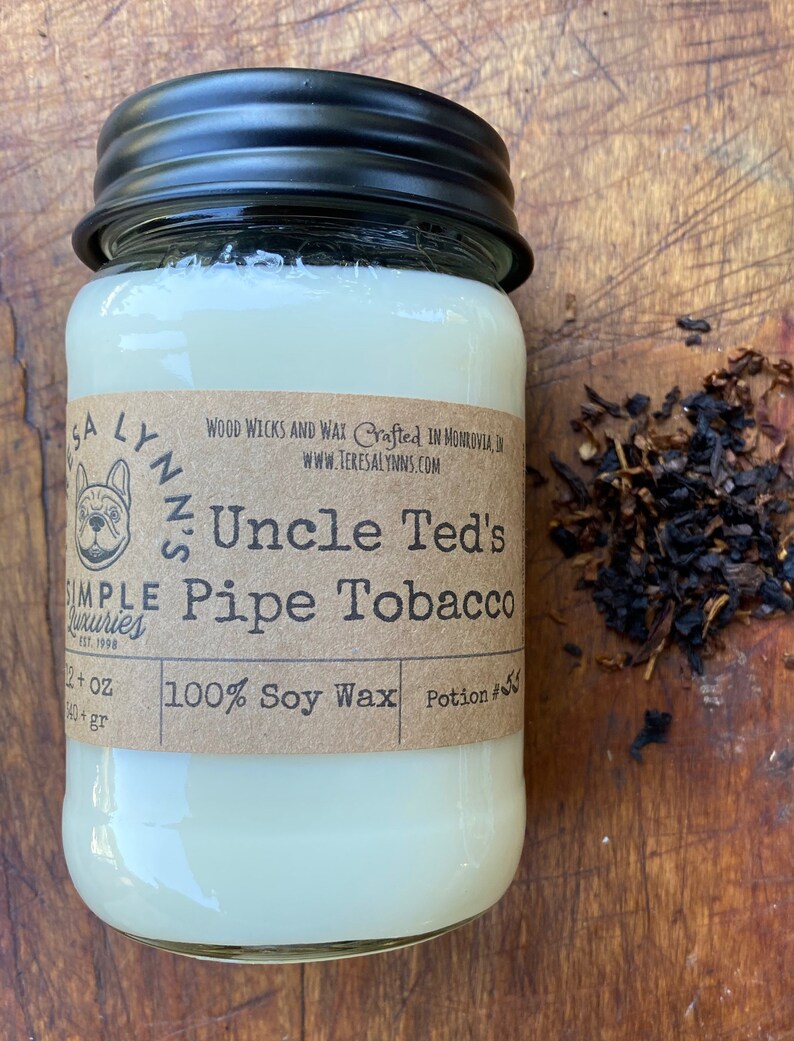 May include: A glass jar candle with a black lid. The label is brown with white text that reads "Uncle Ted's Pipe Tobacco" and "100% Soy Wax". There is a small illustration of a dog on the label.