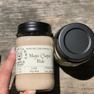 A hand holds a jar candle labeled Magic Carpet Ride with a wood table in the background. Label reads 100% Soy Wax Musk, Amber, Oud. Teresas hand is in frame on the jar.