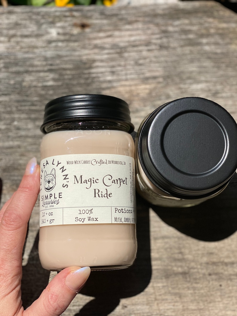 A hand holds a jar candle labeled Magic Carpet Ride with a wood table in the background. Label reads 100% Soy Wax Musk, Amber, Oud. Teresas hand is in frame on the jar.