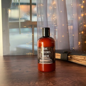 An Amber colored bottle of body wash labeled Witches House, crafted in Monrovia,I n sits on a wooden surface with a grimoire next to it. Behind the bottle is a sheer covered window with twinkly lights and a spider plant.