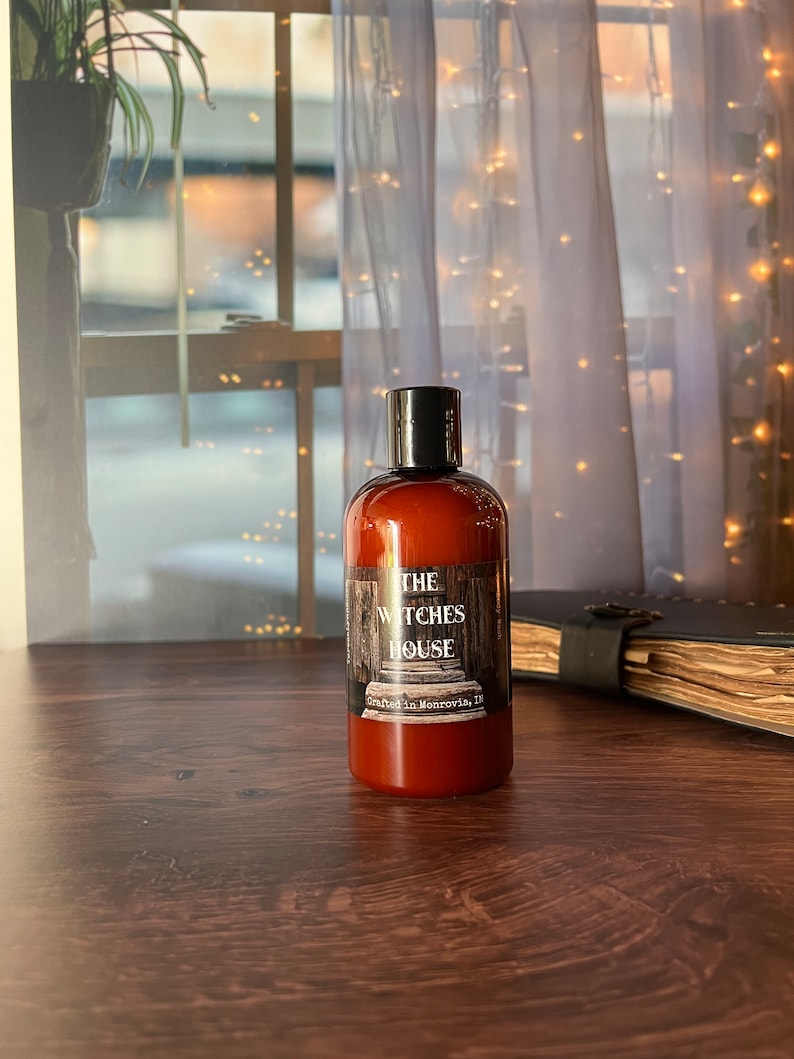 An Amber colored bottle of body wash labeled Witches House, crafted in Monrovia,I n sits on a wooden surface with a grimoire next to it. Behind the bottle is a sheer covered window with twinkly lights and a spider plant.