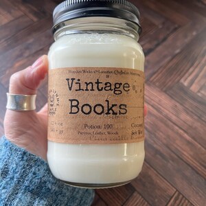 May include: A glass jar candle with a black lid, labeled "Vintage Books." The label is brown with black text, and the candle is filled with white wax. The label also includes the text "12 oz" and "540+ gr."