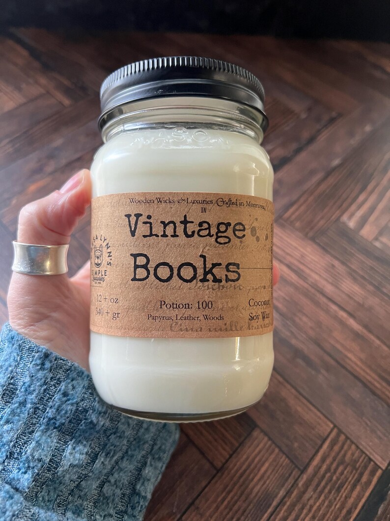 May include: A glass jar candle with a black lid, labeled "Vintage Books." The label is brown with black text, and the candle is filled with white wax. The label also includes the text "12 oz" and "540+ gr."