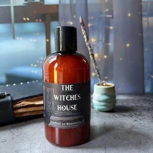 Puede incluir: Una botella cil&iacute;ndrica de pl&aacute;stico &aacute;mbar con tapa de rosca negra. La etiqueta presenta el texto THE WITCHES HOUSE y Crafted in Monrovia, IN. La botella est&aacute; llena de un l&iacute;quido oscuro y tiene una etiqueta rectangular oscura con letras blancas.