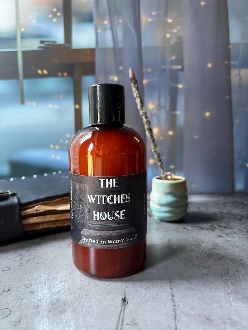 Puede incluir: Una botella cil&iacute;ndrica de pl&aacute;stico &aacute;mbar con tapa de rosca negra. La etiqueta presenta el texto THE WITCHES HOUSE y Crafted in Monrovia, IN. La botella est&aacute; llena de un l&iacute;quido oscuro y tiene una etiqueta rectangular oscura con letras blancas.