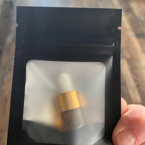 Op de afbeelding: Een klein amberkleurig glazen flesje met een goudkleurige dop en een witte druppelaar, verpakt in een zwart hersluitbaar zakje met een doorzichtig, mat rechthoekig venster aan de voorzijde om de inhoud van de verpakking te tonen.