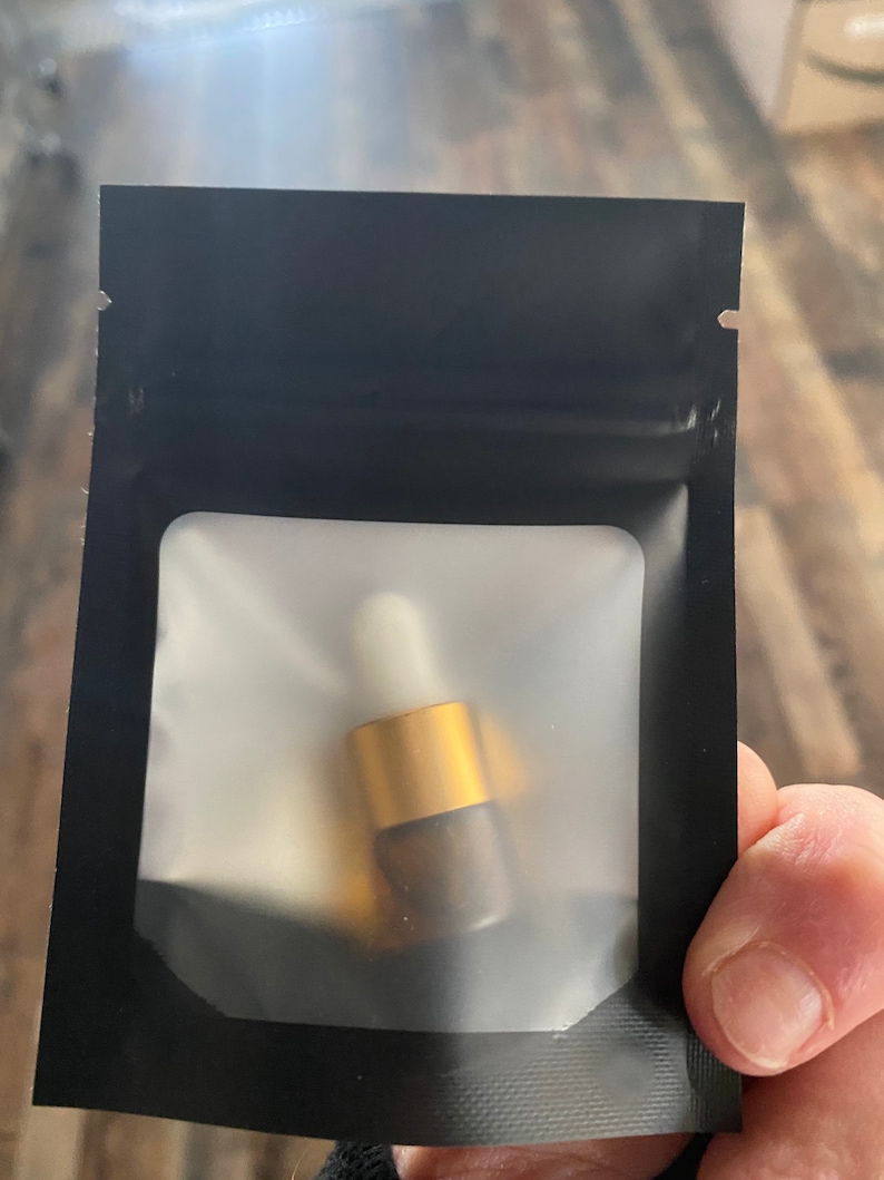 Op de afbeelding: Een klein amberkleurig glazen flesje met een goudkleurige dop en een witte druppelaar, verpakt in een zwart hersluitbaar zakje met een doorzichtig, mat rechthoekig venster aan de voorzijde om de inhoud van de verpakking te tonen.