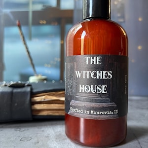 Puede incluir: Una botella cil&iacute;ndrica de pl&aacute;stico &aacute;mbar con tapa negra que contiene gel de ba&ntilde;o. La etiqueta presenta un gr&aacute;fico oscuro de una columna de piedra y el texto: THE WITCHES HOUSE, Crafted in Monrovia, IN, y Body Wash impreso en el lateral.