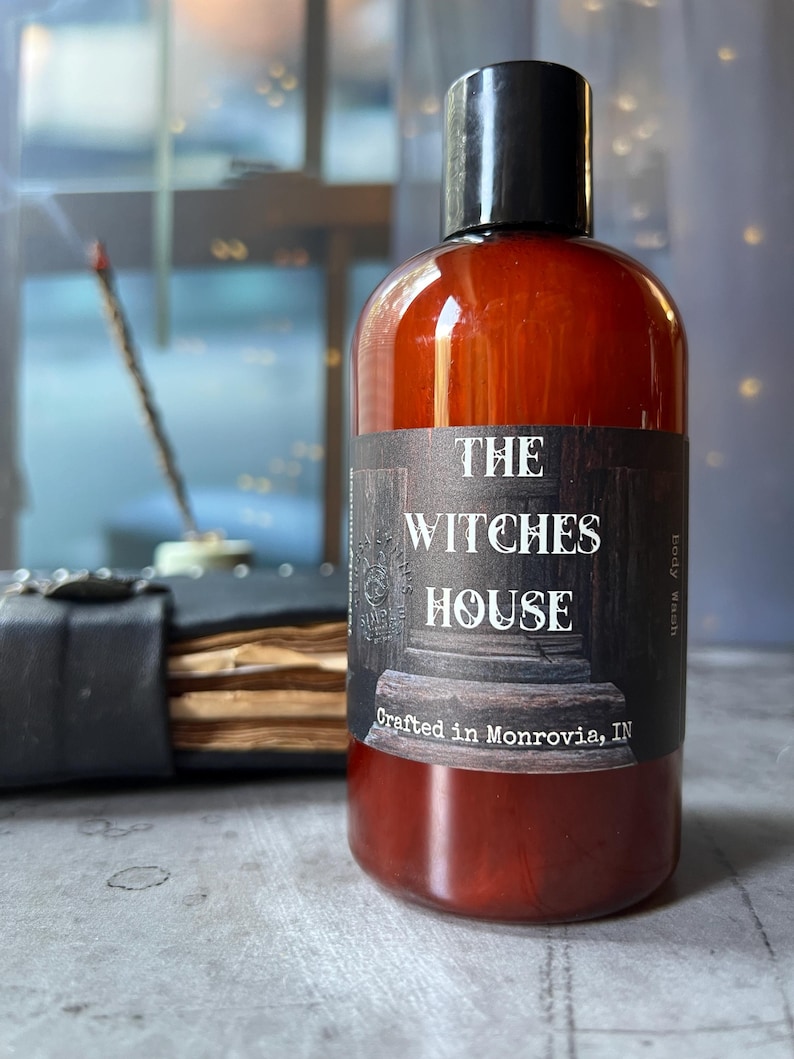 Puede incluir: Una botella cil&iacute;ndrica de pl&aacute;stico &aacute;mbar con tapa negra que contiene gel de ba&ntilde;o. La etiqueta presenta un gr&aacute;fico oscuro de una columna de piedra y el texto: THE WITCHES HOUSE, Crafted in Monrovia, IN, y Body Wash impreso en el lateral.