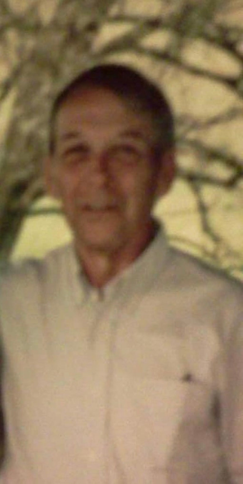 May include: A man wearing a light beige button-down shirt. He is smiling and looking at the camera.
