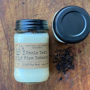 May include: A white soy wax candle in a clear glass jar with a black lid. The candle is labeled "Uncle Ted's Pipe Tobacco" and is made with 100% soy wax. The candle is surrounded by loose tobacco leaves.