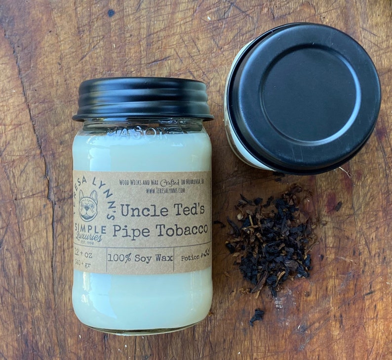May include: A white soy wax candle in a clear glass jar with a black lid. The candle is labeled "Uncle Ted's Pipe Tobacco" and is made with 100% soy wax. The candle is surrounded by loose tobacco leaves.