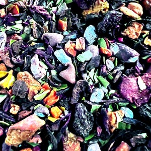 May include: A loose leaf herbal tea blend featuring a vibrant mix of dried dark purple berries, bright orange fruit pieces, small green herbal leaves, and scattered light blue and purple floral petals, creating a textured and colorful botanical mixture.