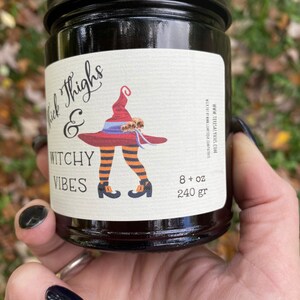 A hand with long black finger nails holds a dark candle jar.  The label reads Thick Thighs & Witchy Vibes.  The graphic is anime inspired witch legs with black and orange striped stockings, topped with red witch hat.  The background is a fall scene.