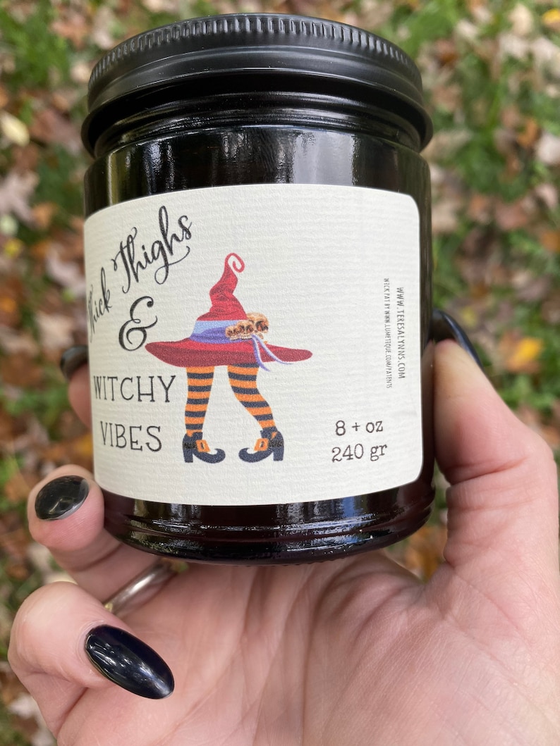 A hand with long black finger nails holds a dark candle jar.  The label reads Thick Thighs & Witchy Vibes.  The graphic is anime inspired witch legs with black and orange striped stockings, topped with red witch hat.  The background is a fall scene.