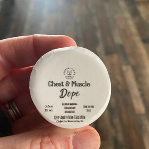 Op de afbeelding: Een kleine witte ronde deksel van een potje met zwarte tekst: Teresa Lynn's Simple Scents, Chest & Muscle Dope, 0.5oz, 15ml, Allergen Warning: Contains Nut Oils, Store in Cool Space en Keep Away From Children.