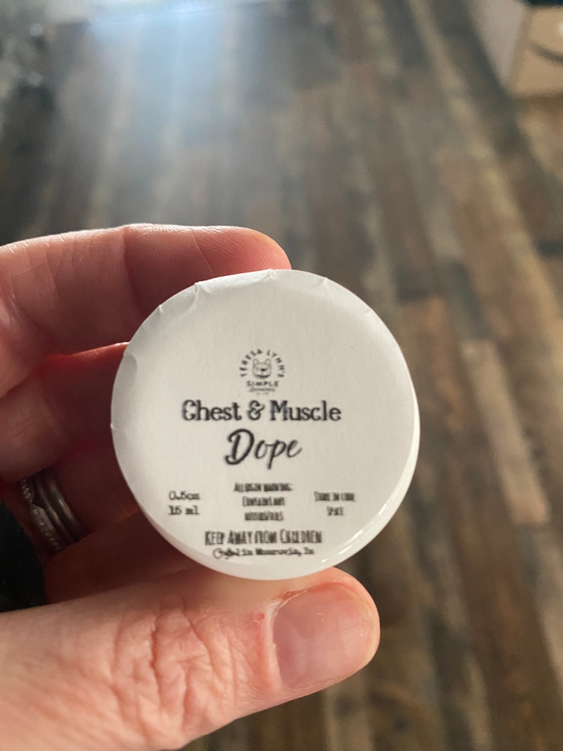 Op de afbeelding: Een kleine witte ronde deksel van een potje met zwarte tekst: Teresa Lynn's Simple Scents, Chest & Muscle Dope, 0.5oz, 15ml, Allergen Warning: Contains Nut Oils, Store in Cool Space en Keep Away From Children.