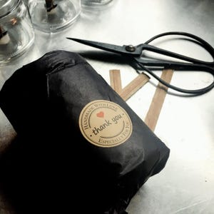 A candle jar wrapped in black tissue paper with a circular Kraft   paper sticker.  Next to the wrapped candle are wooden candle wicks and vintage scissors on a graphite table.