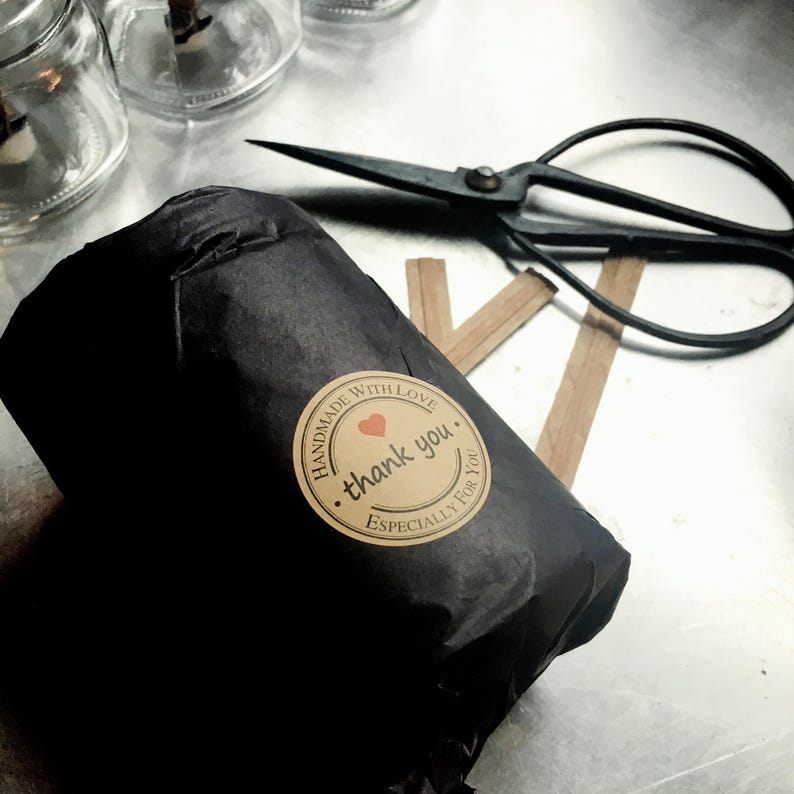A candle jar wrapped in black tissue paper with a circular Kraft   paper sticker.  Next to the wrapped candle are wooden candle wicks and vintage scissors on a graphite table.