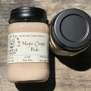 A hand holds a jar candle labeled Magic Carpet Ride with a wood table in the background. Label reads 100% Soy Wax Musk, Amber, Oud