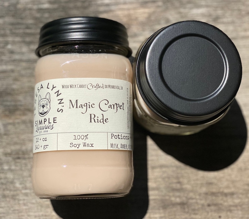 A hand holds a jar candle labeled Magic Carpet Ride with a wood table in the background. Label reads 100% Soy Wax Musk, Amber, Oud