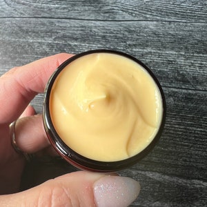 Op de afbeelding: Een klein, rond potje gevuld met lichtgele cr&egrave;me. De cr&egrave;me heeft een gladde, gewervelde textuur. Het potje is zwart met een donkerrode rand. De achtergrond is een donker houten oppervlak.