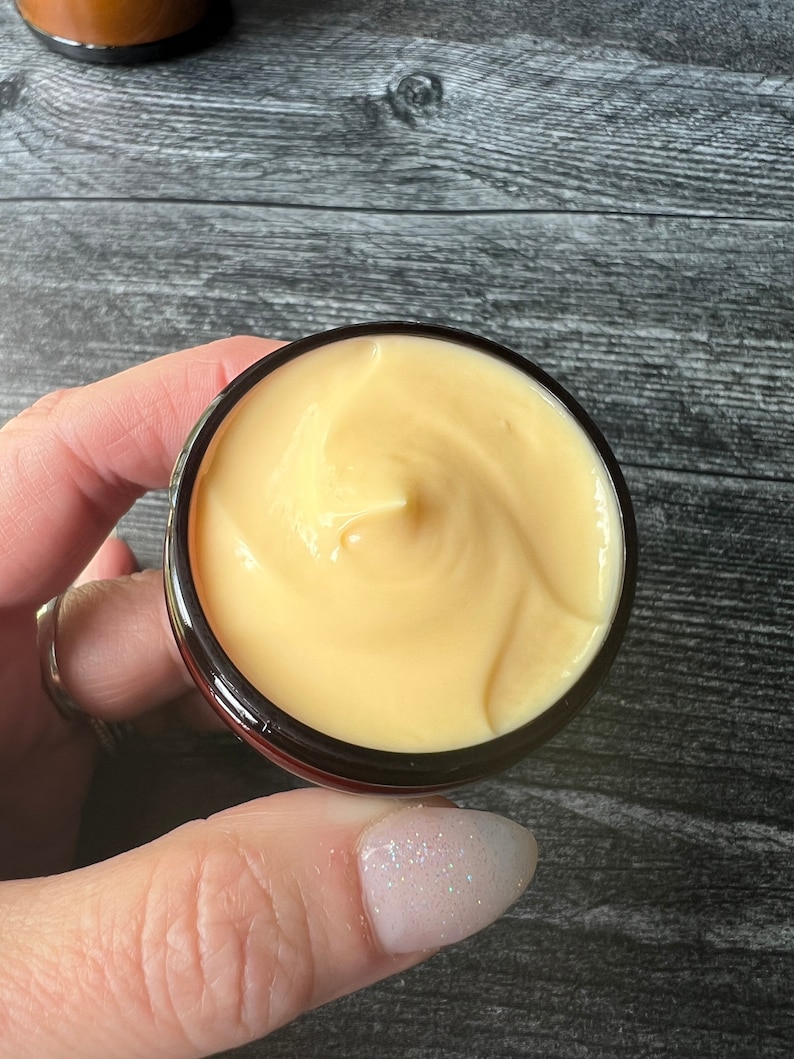 Op de afbeelding: Een klein, rond potje gevuld met lichtgele cr&egrave;me. De cr&egrave;me heeft een gladde, gewervelde textuur. Het potje is zwart met een donkerrode rand. De achtergrond is een donker houten oppervlak.