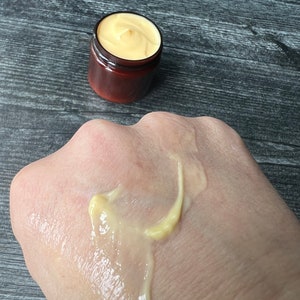 Op de afbeelding: Een klein, bruin potje gevuld met een gele cr&egrave;me, en een kleine hoeveelheid van de cr&egrave;me zit op de hand van een persoon. De cr&egrave;me heeft een gladde, dikke consistentie. Het potje heeft een donkerbruine deksel.