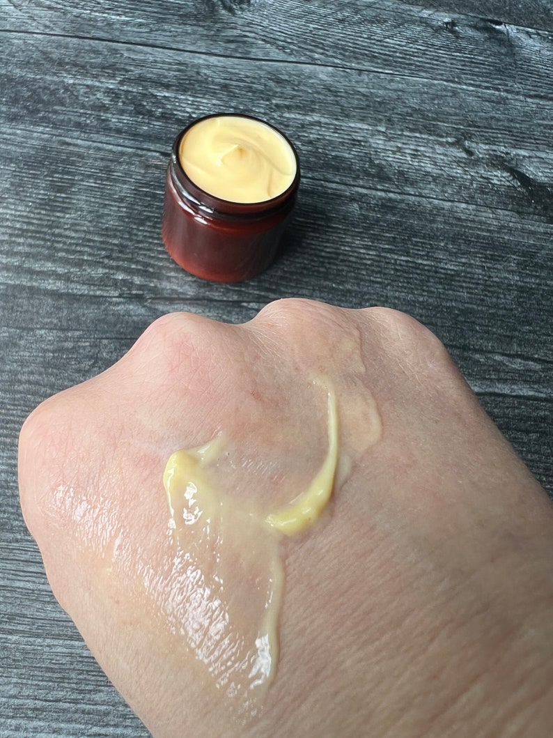 Op de afbeelding: Een klein, bruin potje gevuld met een gele cr&egrave;me, en een kleine hoeveelheid van de cr&egrave;me zit op de hand van een persoon. De cr&egrave;me heeft een gladde, dikke consistentie. Het potje heeft een donkerbruine deksel.