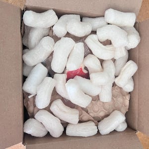 May include: An open cardboard box filled with white packing peanuts. The peanuts are in various curved shapes and fill the box, with a glimpse of the contents beneath. The box is brown.