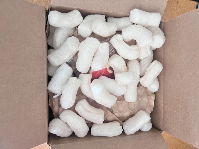 May include: An open cardboard box filled with white packing peanuts. The peanuts are in various curved shapes and fill the box, with a glimpse of the contents beneath. The box is brown.