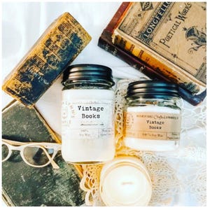 May include: Two glass jars of soy wax candles with black lids, labeled "Vintage Books", sit among antique books and a pair of glasses. The candles are filled with white wax. The image has a vintage aesthetic.
