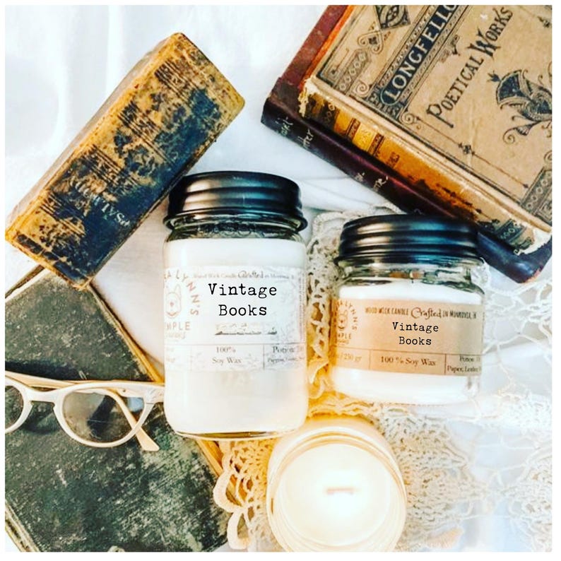 May include: Two glass jars of soy wax candles with black lids, labeled "Vintage Books", sit among antique books and a pair of glasses. The candles are filled with white wax. The image has a vintage aesthetic.