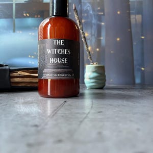 Puede incluir: Una botella cil&iacute;ndrica de pl&aacute;stico &aacute;mbar con tapa negra, que presenta una etiqueta negra con texto blanco que dice THE WITCHES HOUSE y Crafted in Monrovia, IN. La botella contiene un l&iacute;quido oscuro y est&aacute; colocada sobre una superficie gris.